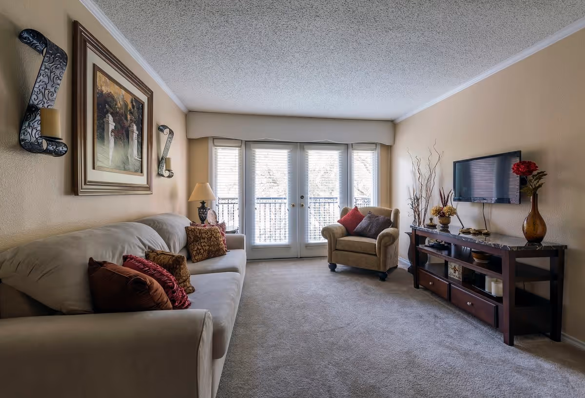 A cozy living room with a beige sofa adorned with multiple decorative pillows on the left, a beige armchair with pillows near the center, and a dark wooden TV stand with a flat-screen TV mounted on the wall. The room has beige walls, a carpeted floor, and large glass doors with blinds leading to a balcony or outdoor area. Wall decorations include a framed painting and two candle holders.