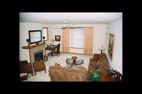 A cozy living room with beige walls and carpet, featuring a brown sofa and loveseat, a round glass coffee table, a fireplace with a mantle holding decorative items, a wooden desk with a chair near a window with beige curtains, and a ceiling fan overhead.