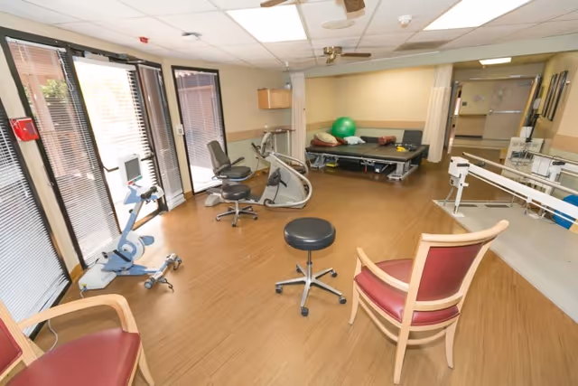 Bright physical therapy/exercise room with exercise bikes, a treatment table, parallel bars and several chairs on a wood floor.