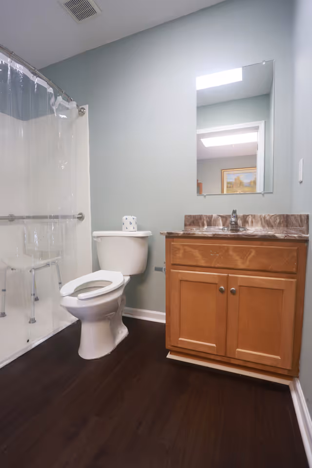 A clean bathroom with a toilet, wooden vanity with a marble countertop and sink, a large mirror above the sink, and a shower area with a clear plastic curtain and a white shower chair inside.