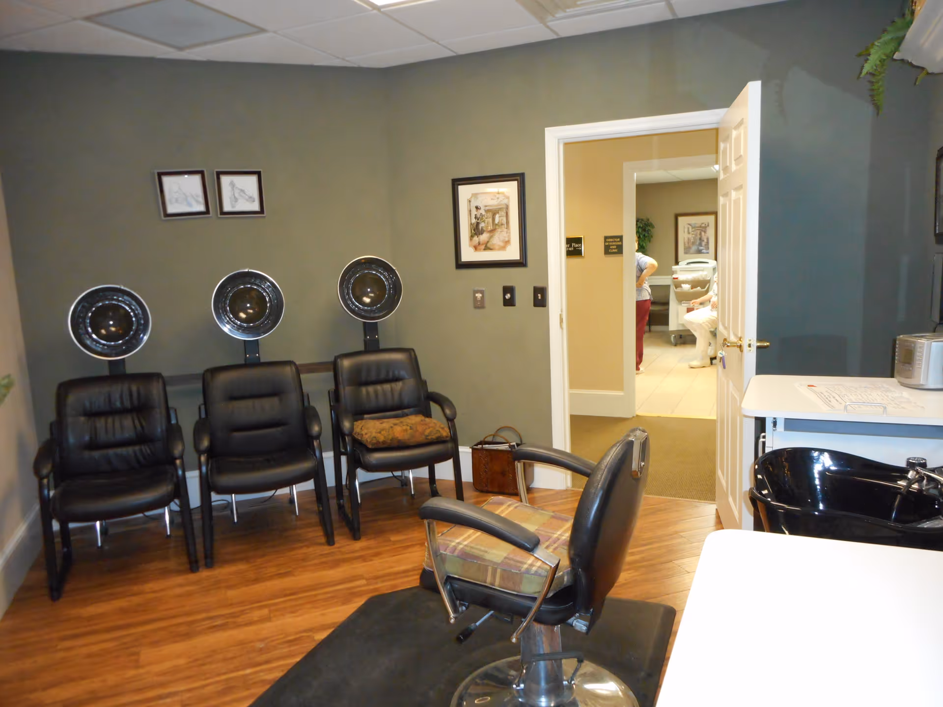 Interior of a hair salon area in a senior living facility with three black leather chairs under hair dryers against a green wall, a salon chair with a plaid cushion in the foreground, and a black hair washing sink on the right. An open door reveals a hallway with two people partially visible and framed pictures on the walls.