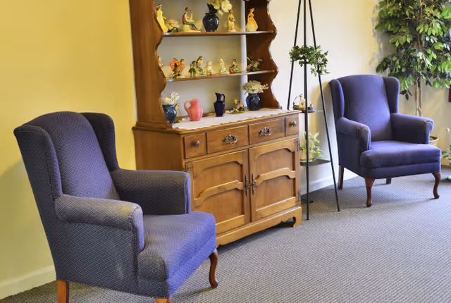 Two purple upholstered armchairs flank a wooden hutch displaying decorative figurines in a carpeted sitting area with plants.