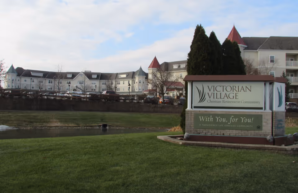 Exterior view of Victorian Village Christian Retirement Community building with a large sign in the foreground that reads 'Victorian Village Christian Retirement Community' and 'With You, for You! A Providence Life Services Community'. The building has multiple floors, peaked roofs, and several windows. There is a grassy area and a small pond in front of the building under a partly cloudy sky.