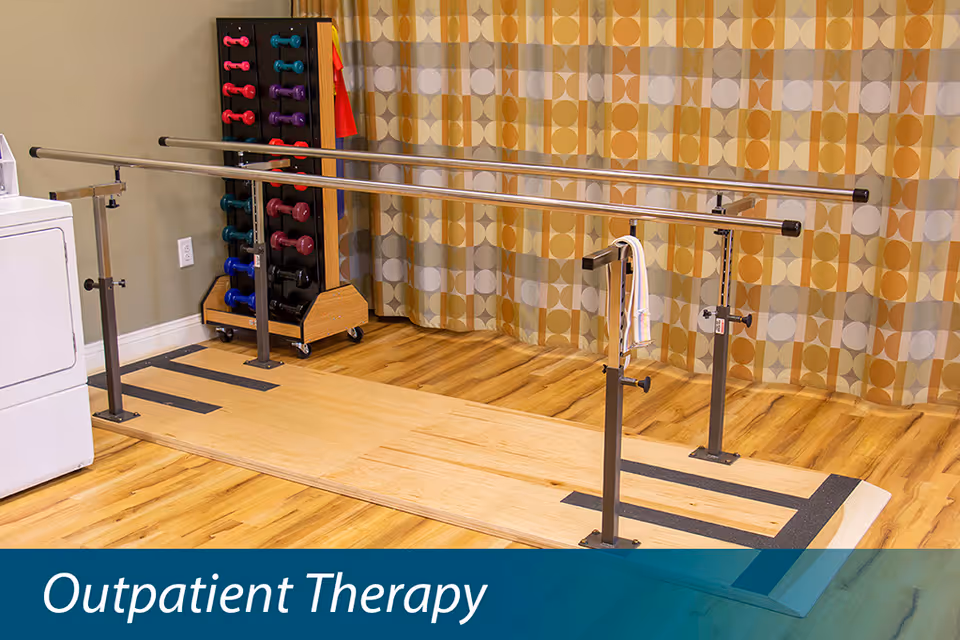 A physical therapy area with parallel bars for walking exercises, a rack of colorful hand weights, and a patterned curtain in the background. The floor is wooden, and there is a white appliance partially visible on the left side.