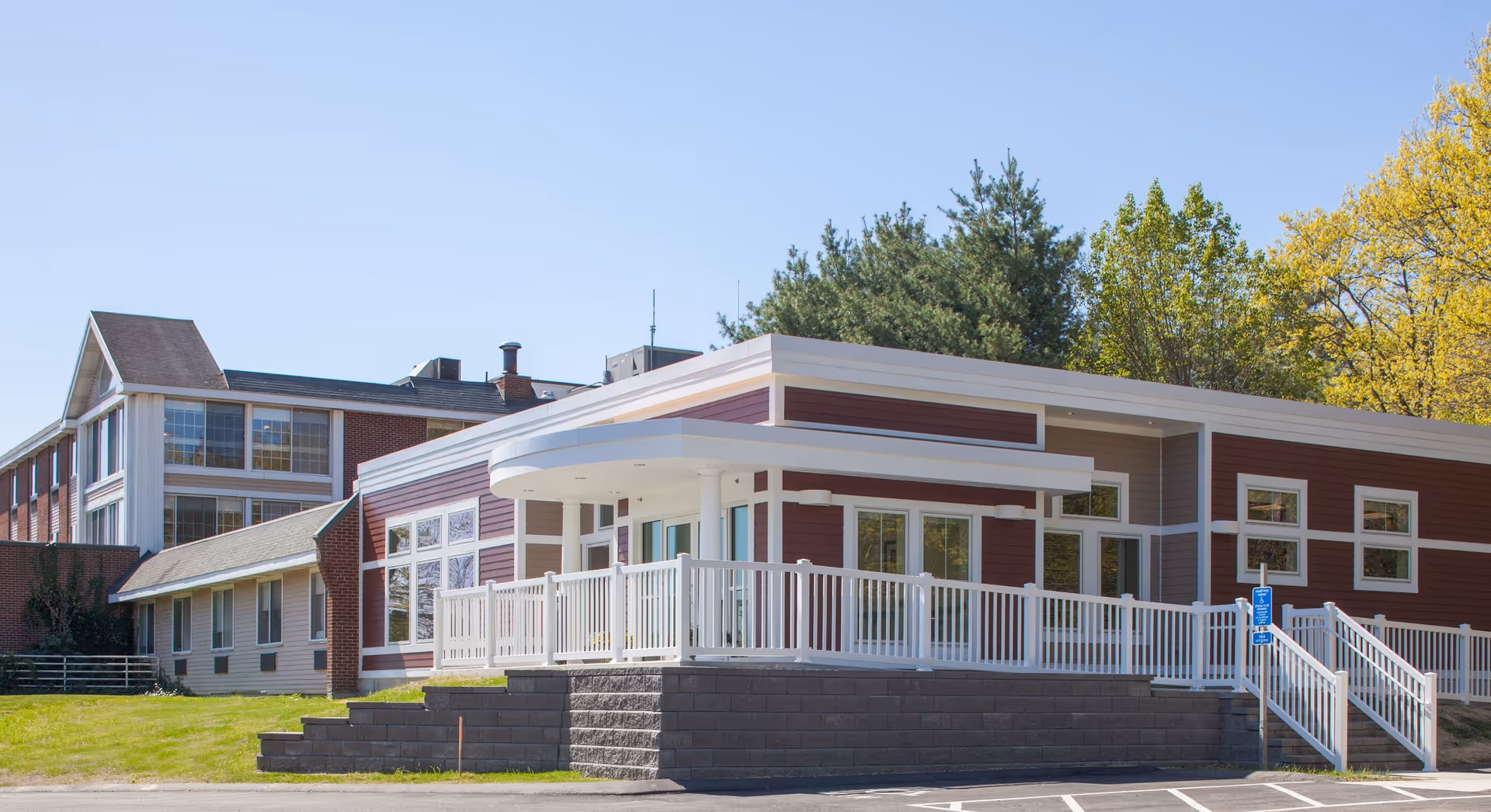 Exterior view of Andover Forest Post Acute Care Center showing a modern building with red and beige siding, white railings, multiple windows, and a ramp leading to the entrance. The building is surrounded by green grass and trees under a clear blue sky.