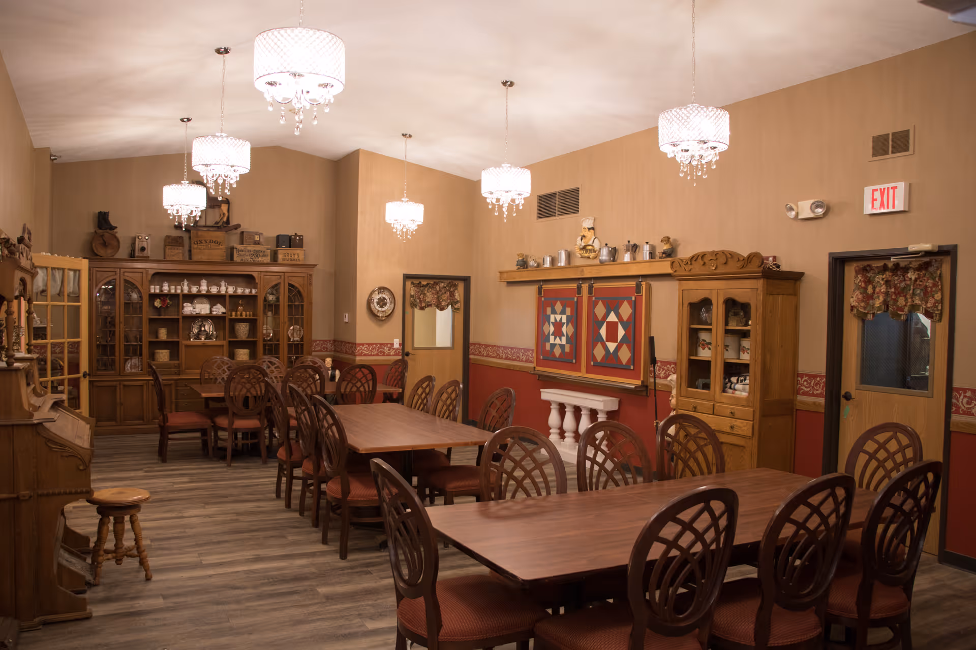 Warmly lit dining room with wooden tables and chairs, chandeliers, and display cabinets with decorative items.