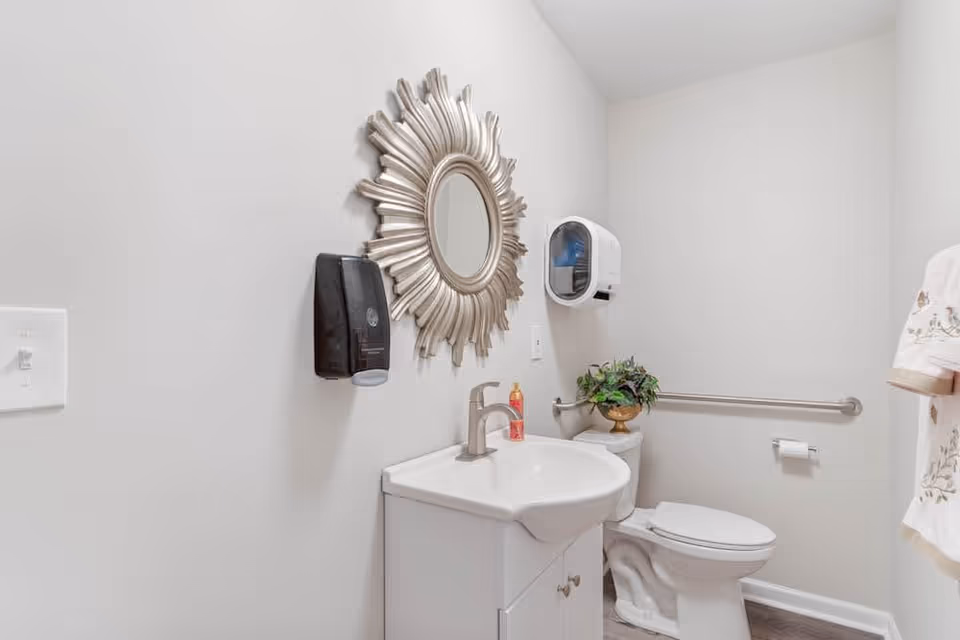A clean and bright bathroom with a white sink and cabinet, a silver faucet, a decorative sunburst mirror above the sink, a black soap dispenser mounted on the wall, a white toilet with a small green plant on the tank, a wall-mounted paper towel dispenser, a grab bar next to the toilet, and a towel hanging on the right side.
