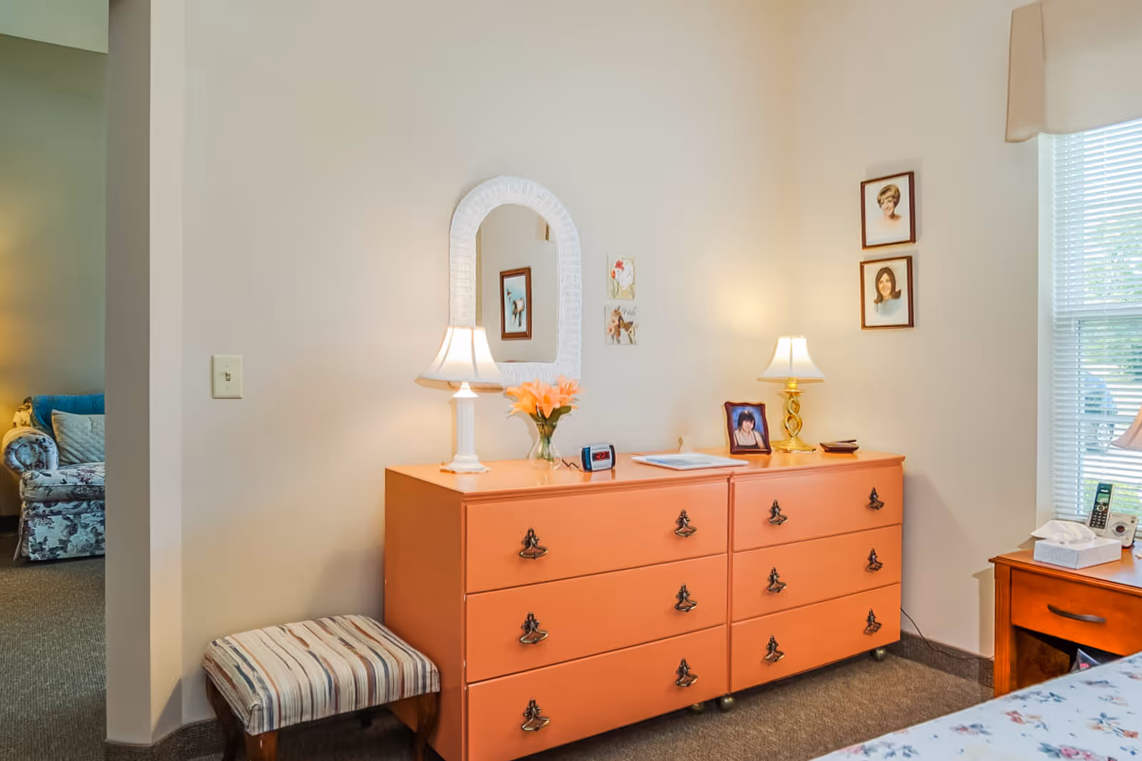 A peach-colored dresser topped with two lamps, framed photos and flowers sits against a bedroom wall next to a window and a striped bench.
