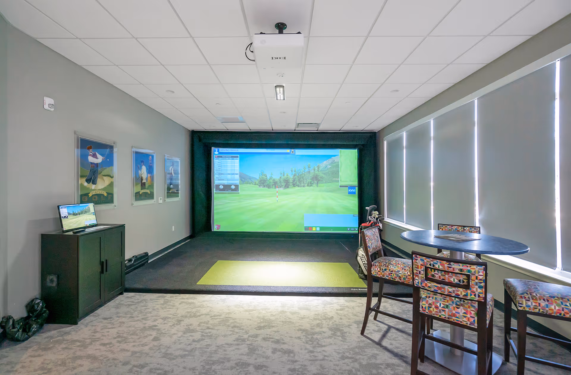 Indoor golf simulator room with a large screen displaying a golf course. The room has a green mat for hitting golf balls, a small round table with three colorful patterned chairs, and a cabinet with a laptop on top. The walls are gray with three framed pictures of golfers, and the windows have closed blinds.