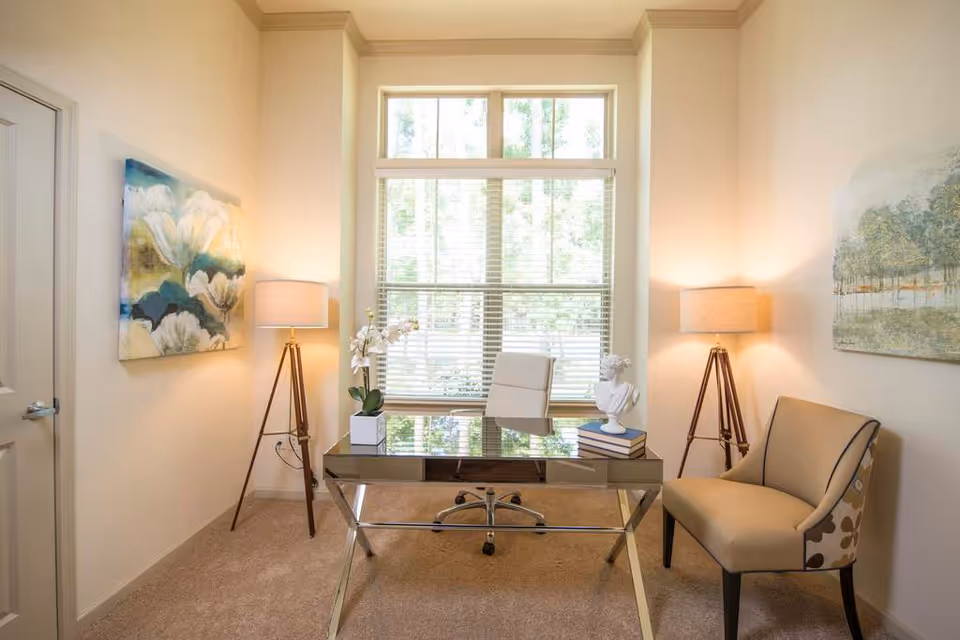 Bright home office with a glass desk, swivel chair, upholstered accent chair, two floor lamps, and a large window with blinds.