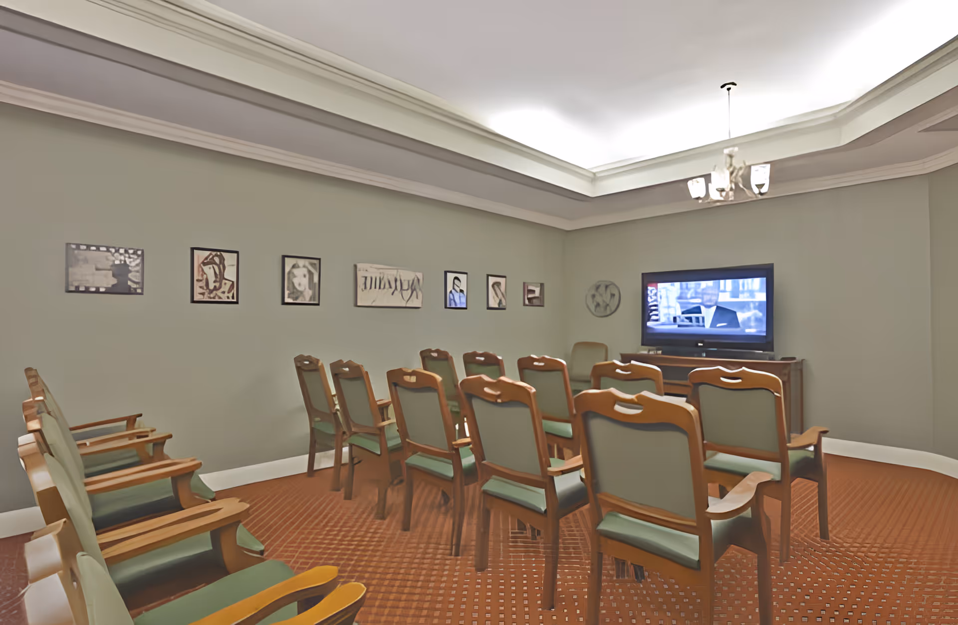 A small theater or media room with rows of wooden chairs with green cushions facing a flat-screen TV mounted on a wooden stand. The walls are painted light green and decorated with framed artwork. The ceiling has recessed lighting and a chandelier.