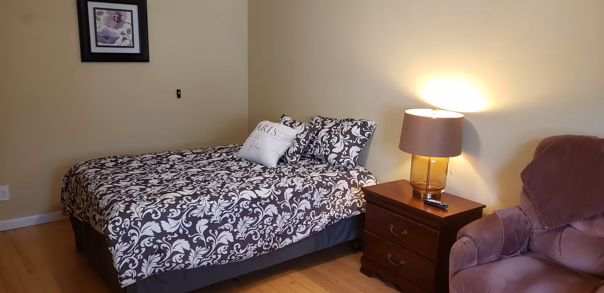 A bedroom with a bed covered in a black and white floral patterned bedspread, two matching pillows, and a decorative pillow with the word 'Paris'. Next to the bed is a wooden nightstand with a lamp and a remote control. A comfortable upholstered armchair is positioned nearby, and a framed floral picture hangs on the beige wall above the bed.