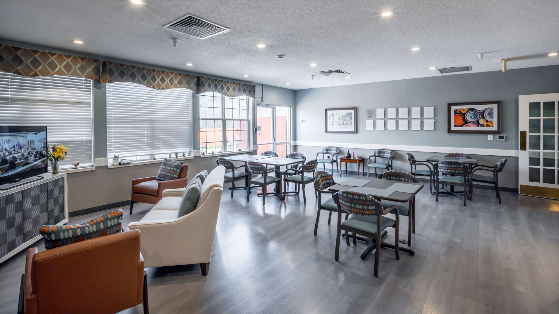 Bright communal dining and lounge area with multiple tables and chairs, sofas, and a television near large windows.