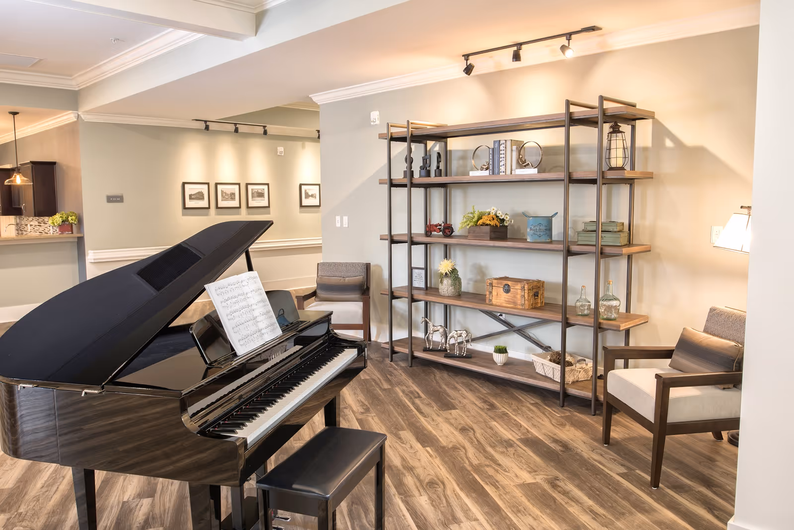 A bright and spacious interior room featuring a black grand piano with sheet music on the stand, a wooden shelving unit with decorative items such as books, plants, and small sculptures, and two cushioned armchairs. The room has wood flooring, soft beige walls, and framed pictures on the far wall.