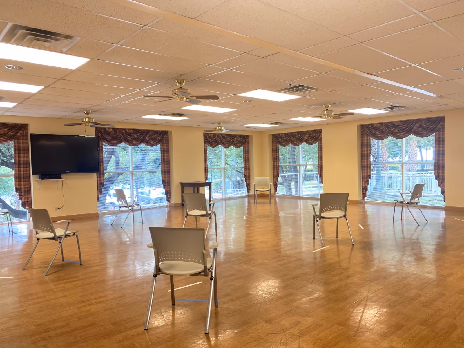Spacious activity room with widely spaced chairs on a wood floor, large windows with valances, ceiling fans, and a wall-mounted TV.