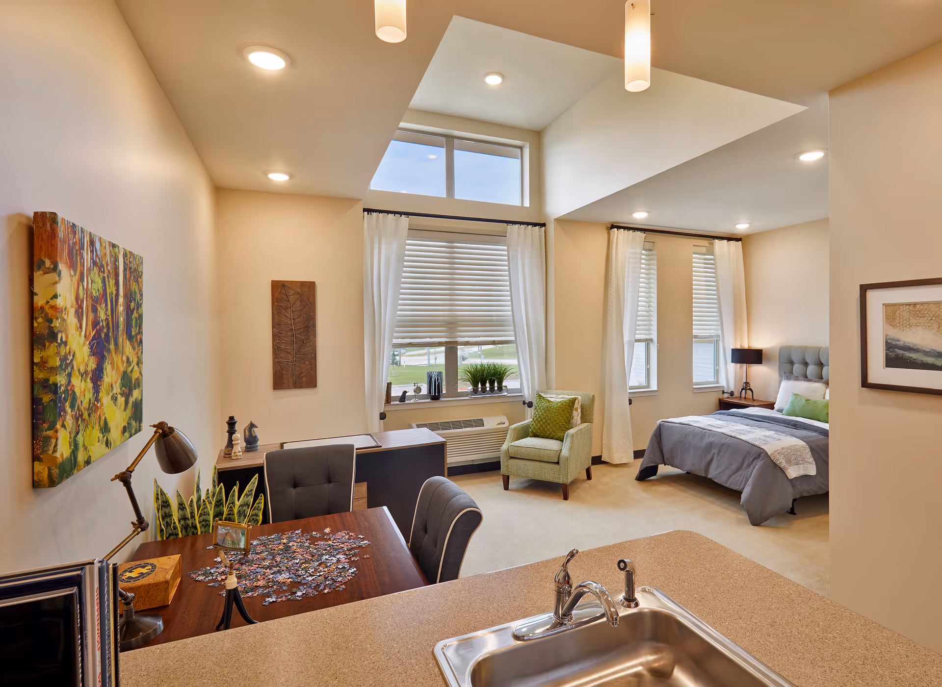 A bright and spacious senior living apartment featuring a cozy bedroom with a bed, nightstand, and lamp, a comfortable armchair by large windows with white curtains, a desk with a chair, and a dining table with chairs and a puzzle in progress. The room has beige walls, recessed lighting, and a kitchen counter with a sink in the foreground.