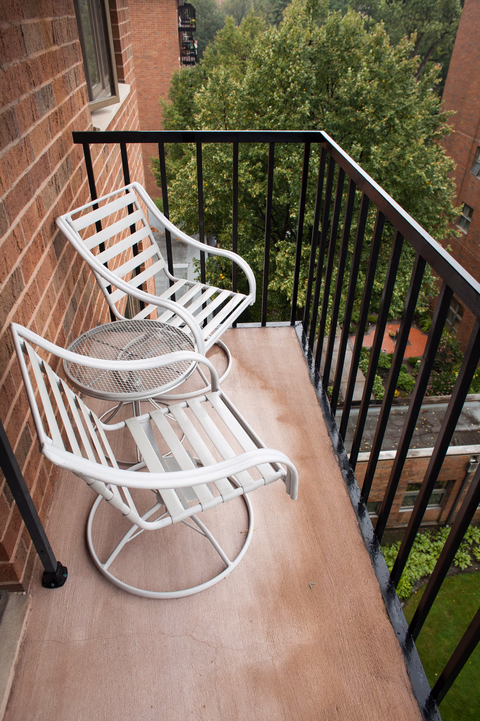 Small outdoor balcony with two white metal chairs and a matching round table. The balcony has a black metal railing and overlooks green trees and parts of a brick building.