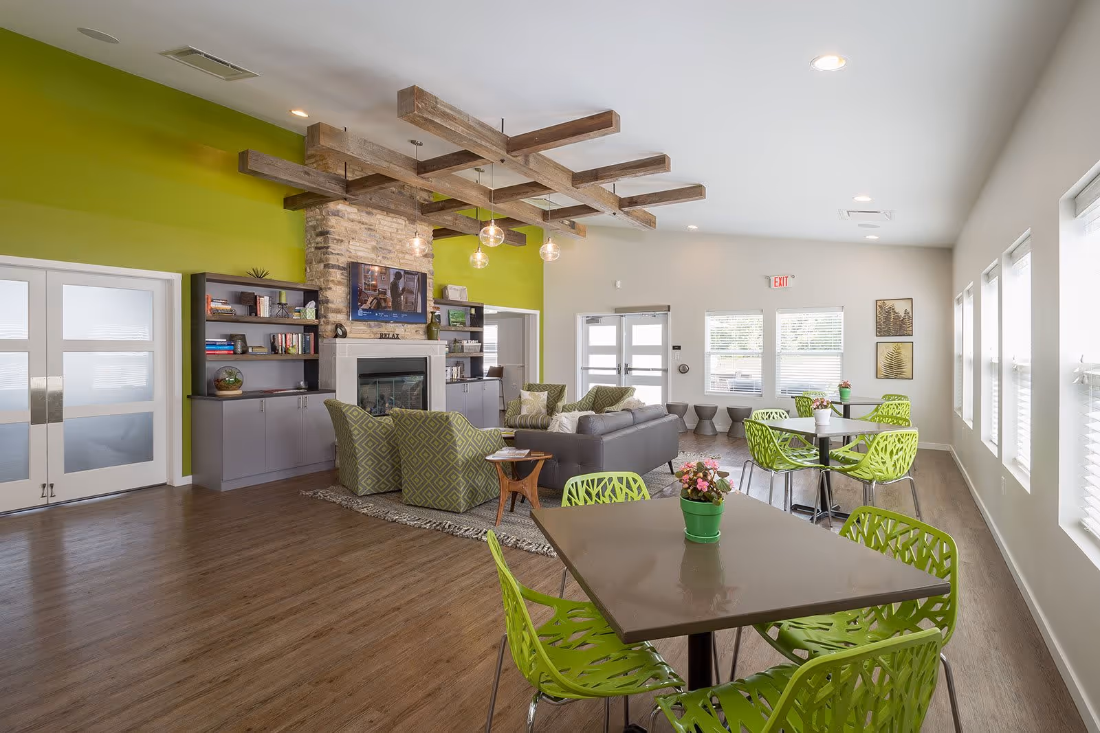 A bright and modern common area with green accent walls and wooden flooring. The room features a cozy seating area with a gray sofa, green patterned armchairs, a stone fireplace with a mounted TV above it, and built-in shelves. There are several tables with green chairs and small potted plants on top, large windows letting in natural light, and decorative wooden beams on the ceiling.