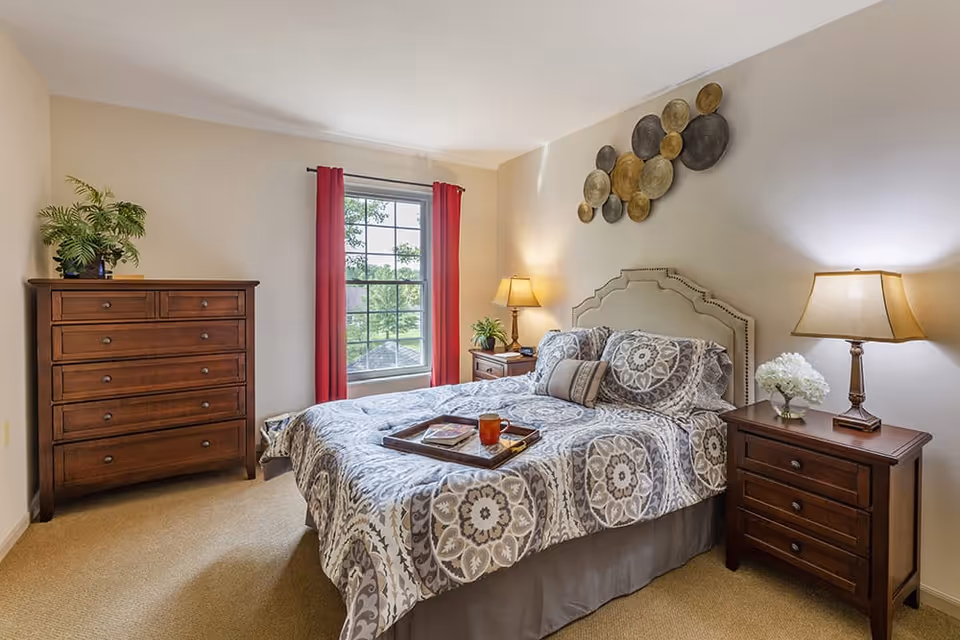 Well-lit bedroom with a double bed dressed in patterned bedding, two nightstands with lamps, a wooden dresser, a window with red curtains, and decorative wall art.