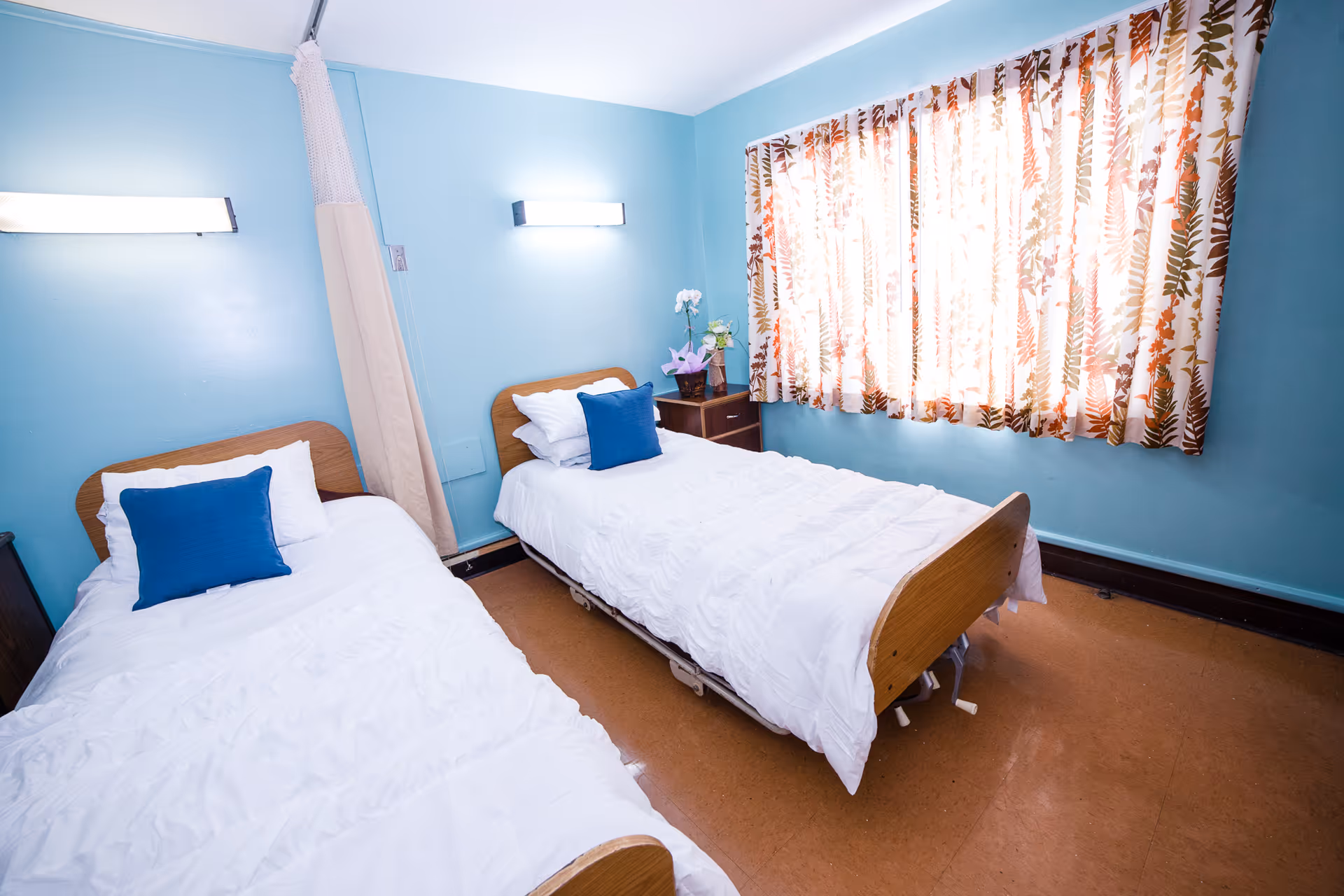 A small bedroom with two single beds, each made with white bedding and a blue pillow. The walls are painted light blue, and there is a window with floral patterned curtains letting in sunlight. A small wooden nightstand with a vase of flowers is placed between the beds. Two wall-mounted lights are above each bed.