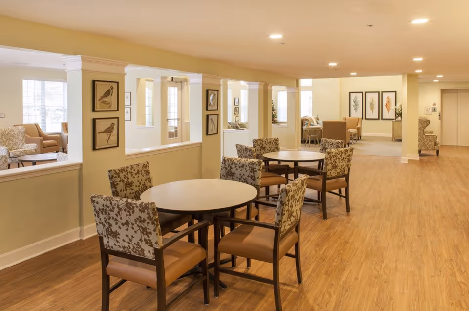 A spacious, well-lit common area in a senior living facility with round tables and cushioned chairs featuring floral upholstery. The room has light yellow walls, wood flooring, and several framed bird and leaf artworks on the walls. There are multiple seating areas with armchairs and a hallway leading to an elevator.