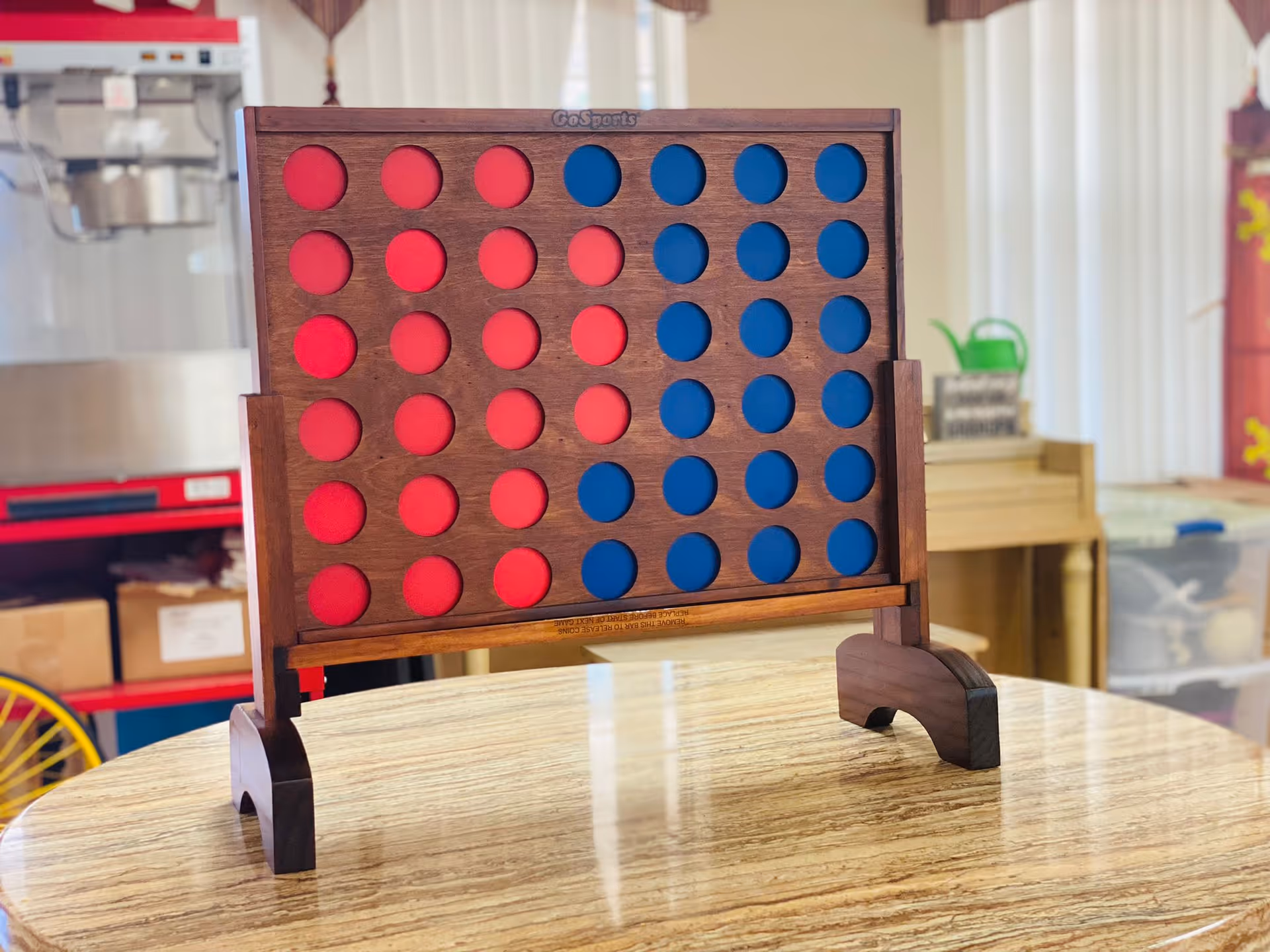 A wooden Connect Four game with red and blue pieces standing on a round wooden table in a communal room.