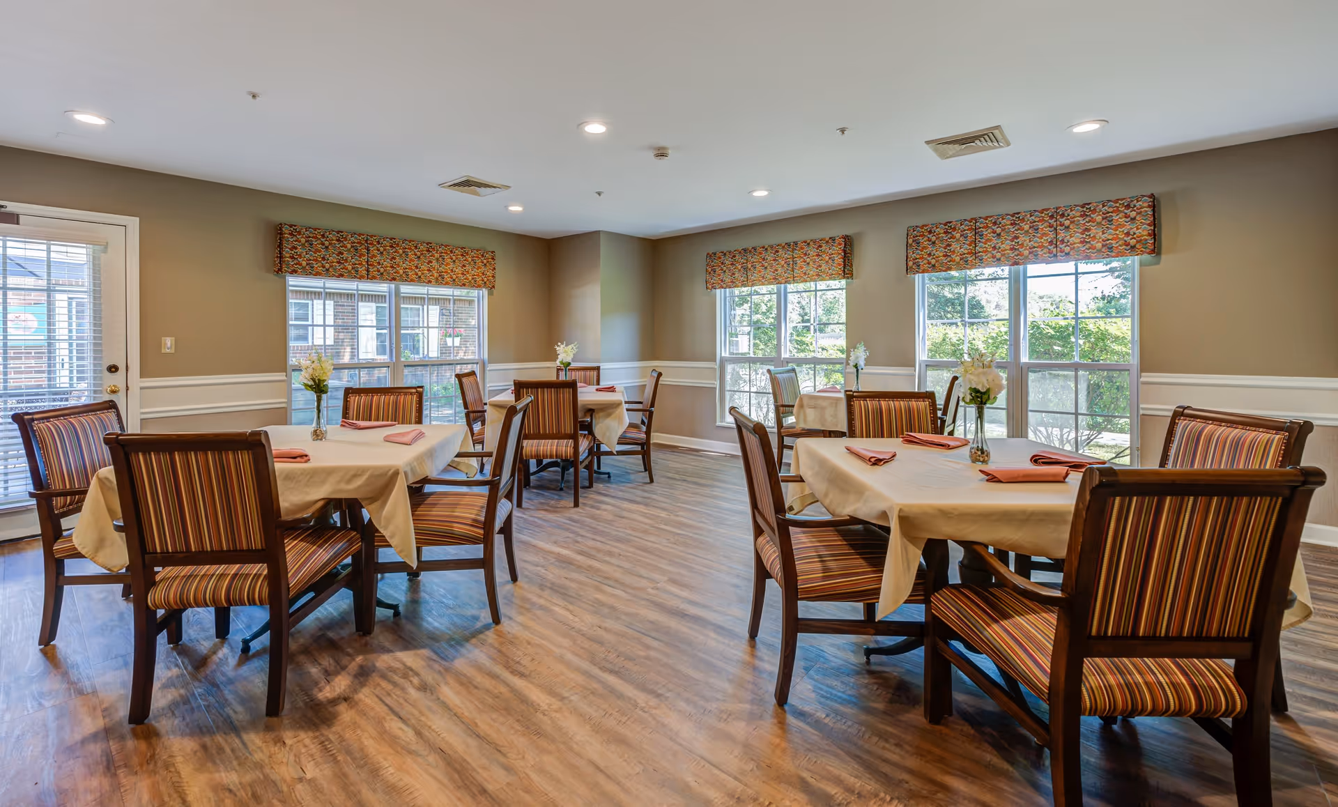 A bright dining room with several tables covered with beige tablecloths and set with folded pink napkins. Each table is surrounded by wooden chairs with striped upholstery. Large windows with patterned valances allow natural light to fill the room, and small flower arrangements are placed on each table. The floor is wood, and the walls are painted beige with white wainscoting.