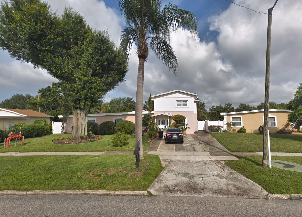 Front exterior view of a two-story residential building with a driveway, a black car parked, a person standing next to the car, a large tree and a palm tree in the front yard, and a cloudy sky.