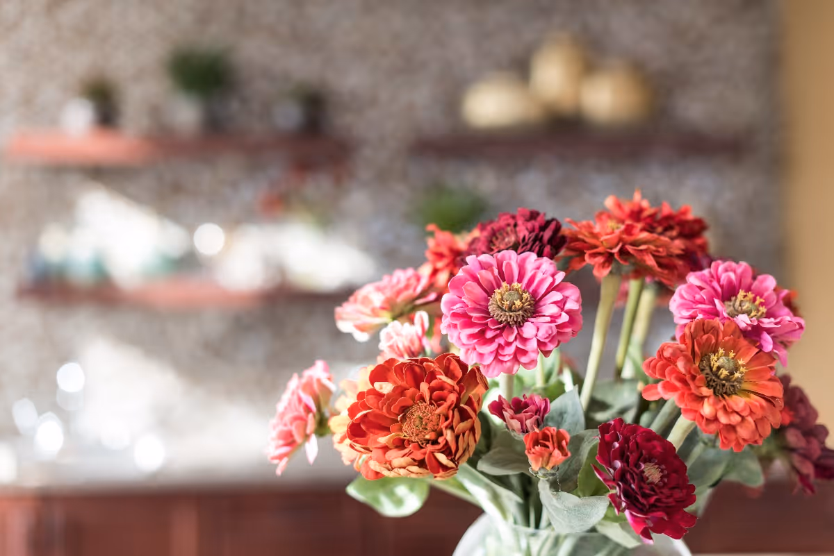 Close-up of a bouquet of vibrant pink, red, and orange flowers in a vase with a softly blurred background featuring shelves and decorative items.