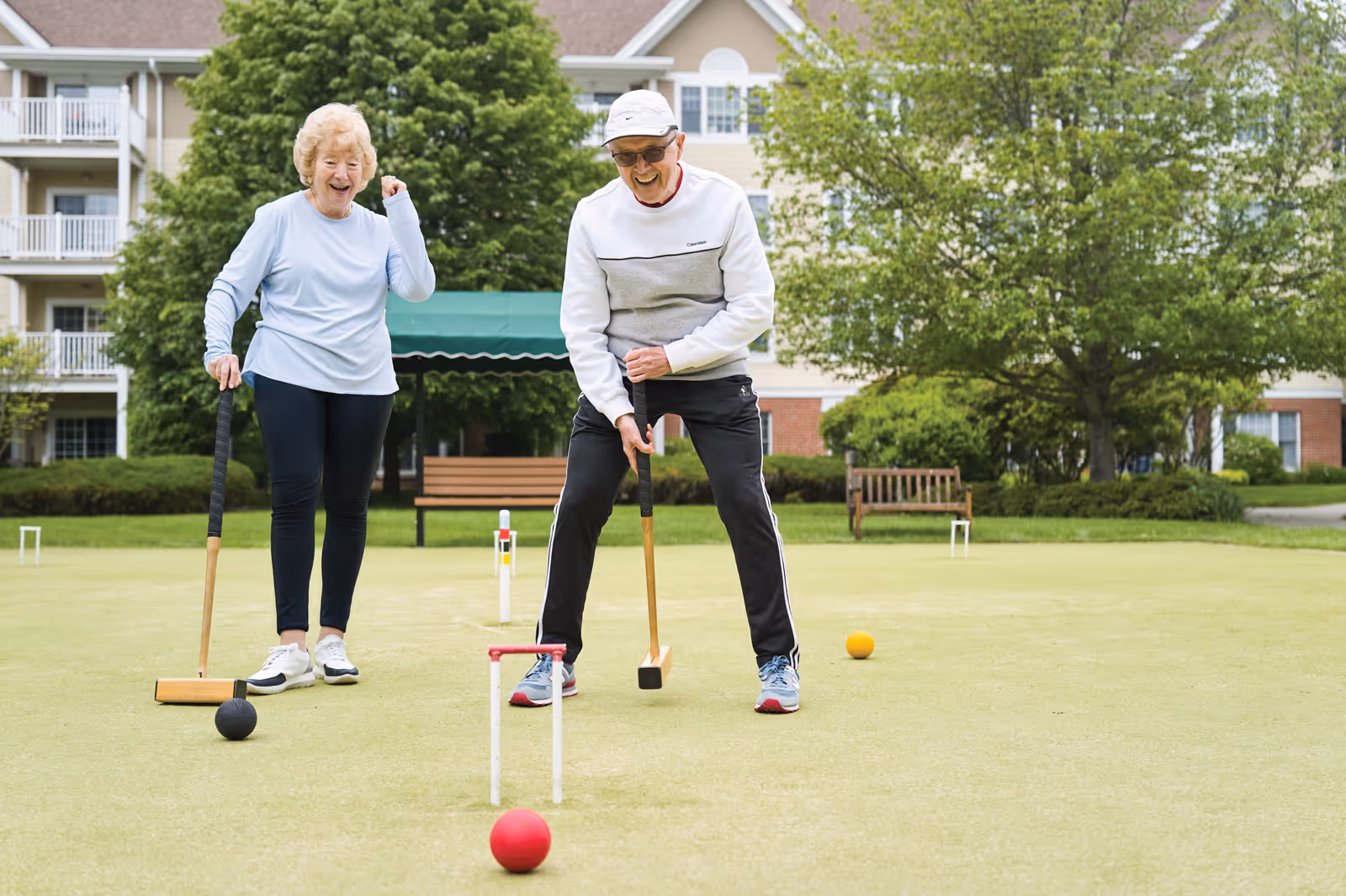Two elderly people playing croquet on a green lawn in front of a residential building with trees and benches in the background. One person is about to hit a red ball with a mallet while the other watches and smiles.