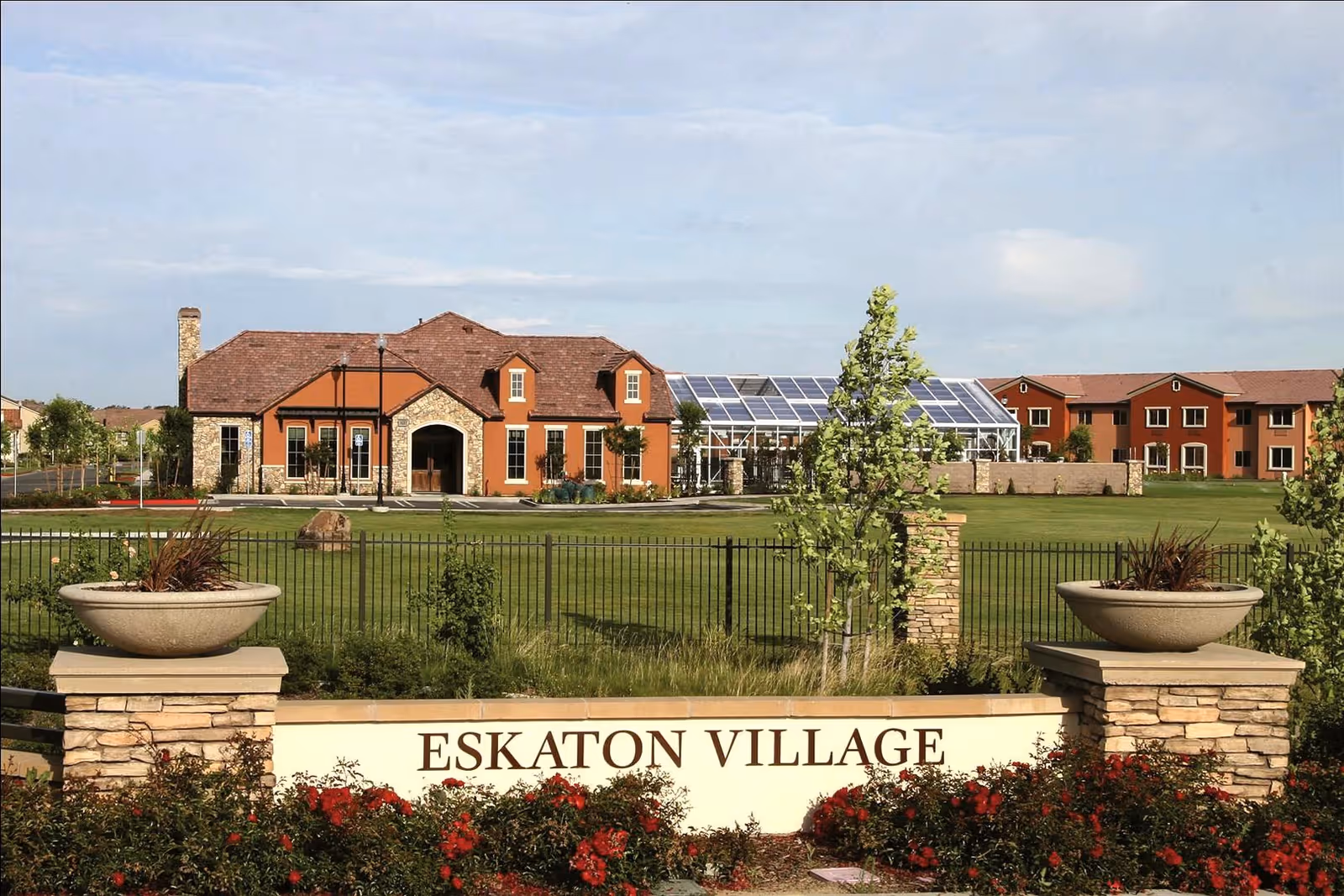 View of Eskaton Village Roseville facility showing a large green lawn with a fence, two stone pillars with planters, and a building with a brown roof and stone facade in the background under a partly cloudy sky.