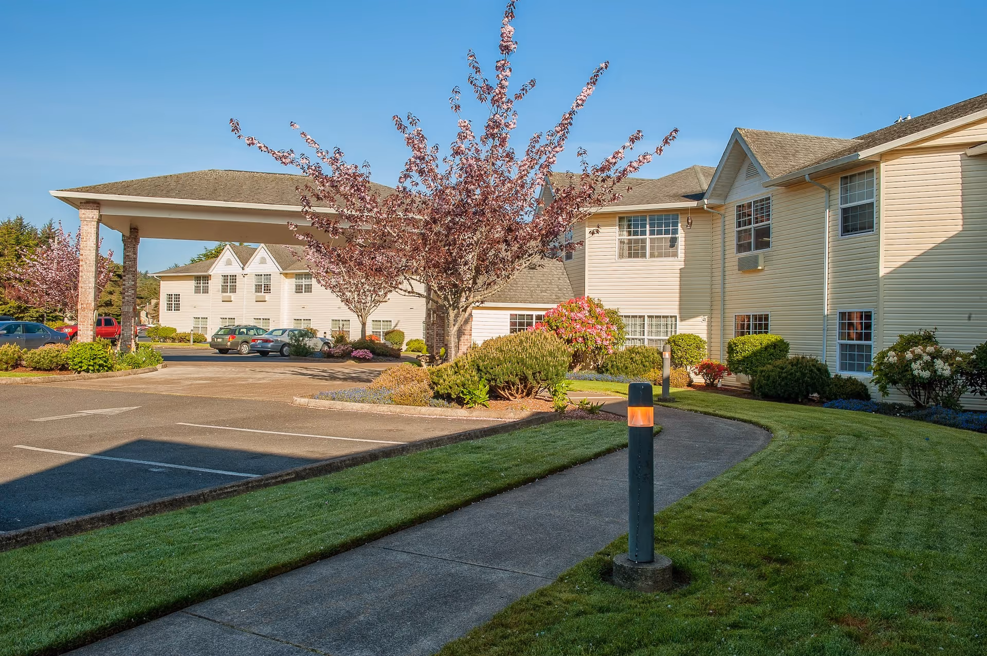 Exterior view of Lakeview Senior Living facility featuring a two-story beige building with multiple windows, a covered entrance supported by brick columns, a paved parking area with several cars, landscaped greenery including blooming trees and bushes, and a clear blue sky.