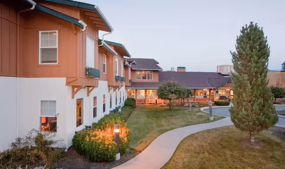 Exterior view of a senior living building with a curved walkway, lawn, trees, and warm interior lights at dusk.