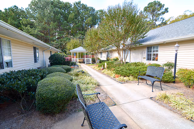 Outdoor courtyard area at a senior living facility with a concrete walkway, black metal benches, manicured bushes, trees, and a white gazebo in the background. Beige single-story buildings with windows flank the walkway on both sides.