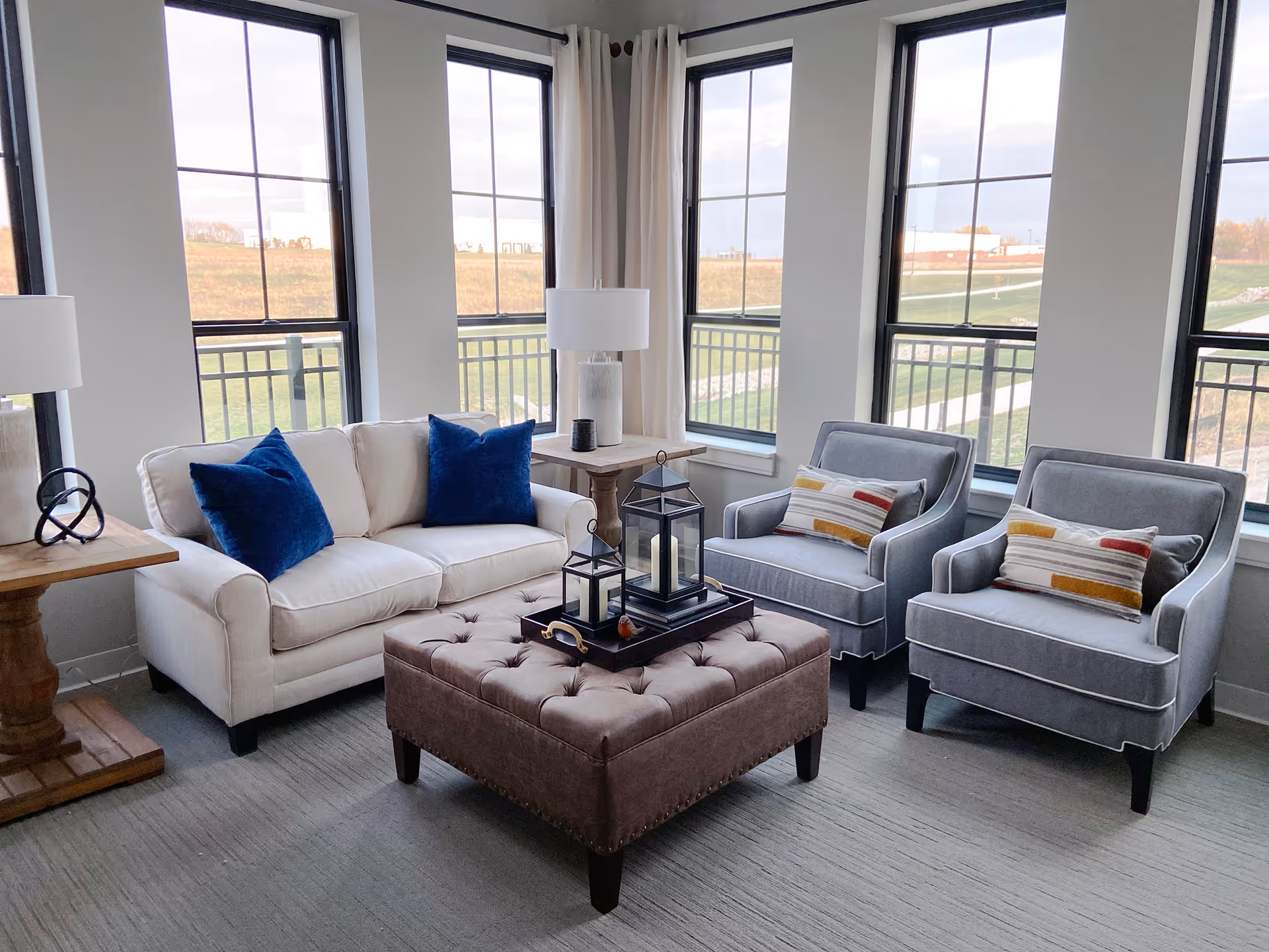 A bright living room with large windows showing an outdoor view. The room features a white sofa with two blue pillows, two gray armchairs with striped cushions, a tufted brown ottoman with decorative lanterns on top, and two wooden side tables each with a white lamp.