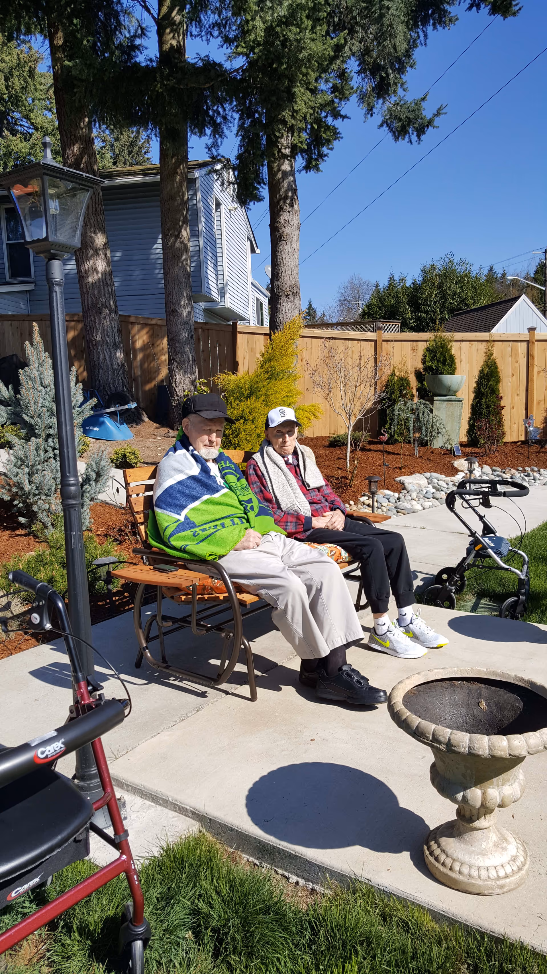 Two elderly adults sitting on a bench in a sunny fenced backyard patio with walkers, landscaping, and a small fire pit nearby.