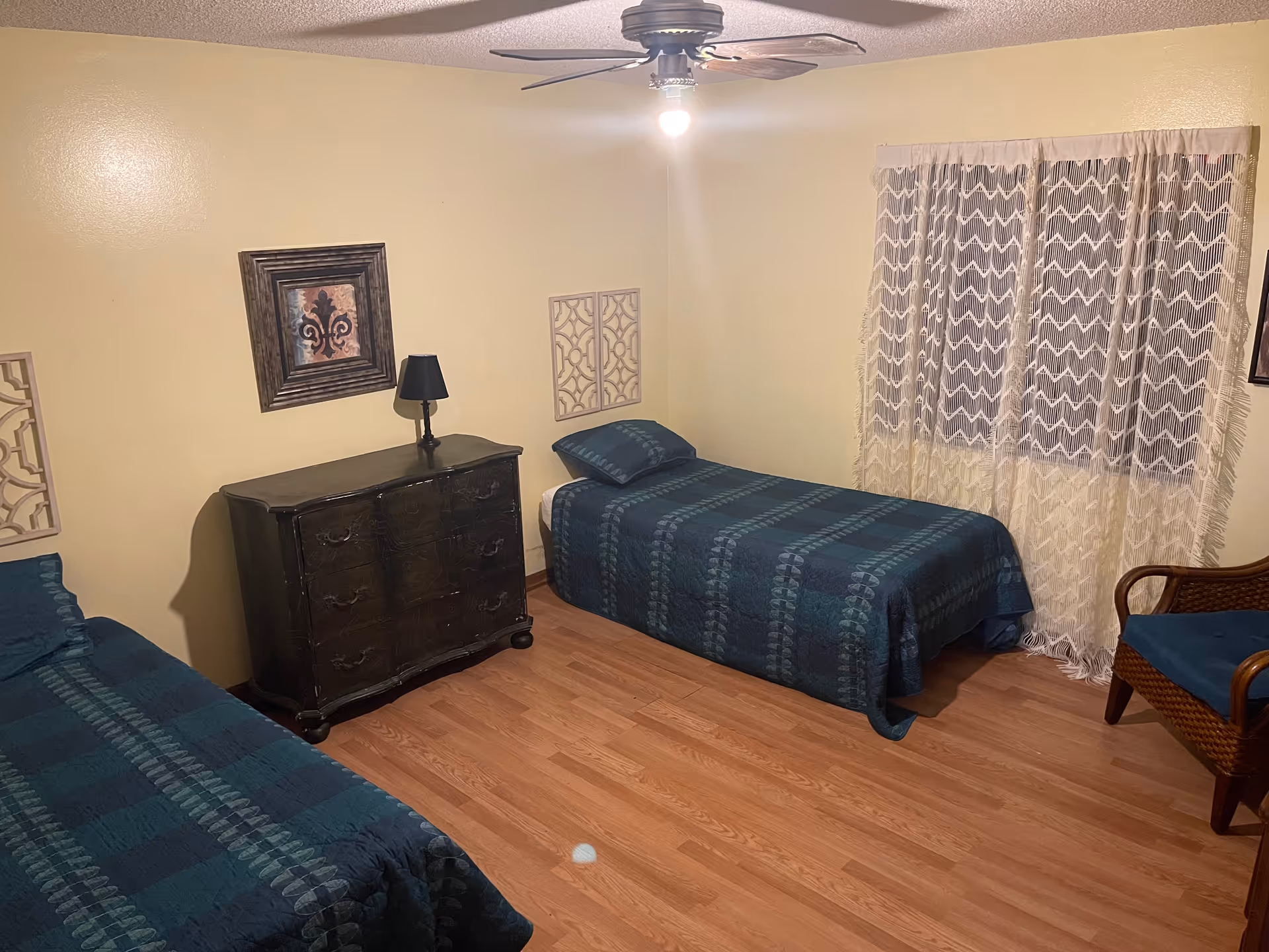 A bedroom with two single beds covered in dark blue patterned bedspreads. There is a dark wooden dresser with a black lamp on top between the beds. A wicker chair with a blue cushion is placed near a window with sheer white curtains. The room has light yellow walls and wooden flooring, with a ceiling fan and light fixture above.
