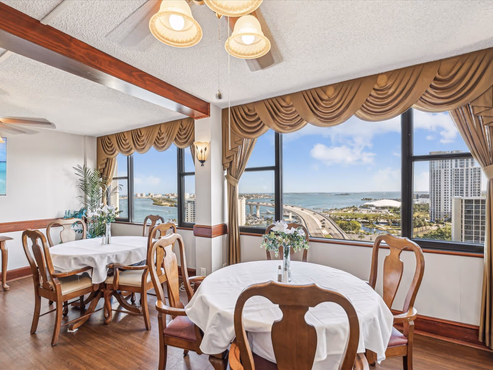 Sunlit dining room with round tables and wooden chairs overlooking a waterfront view through large windows.