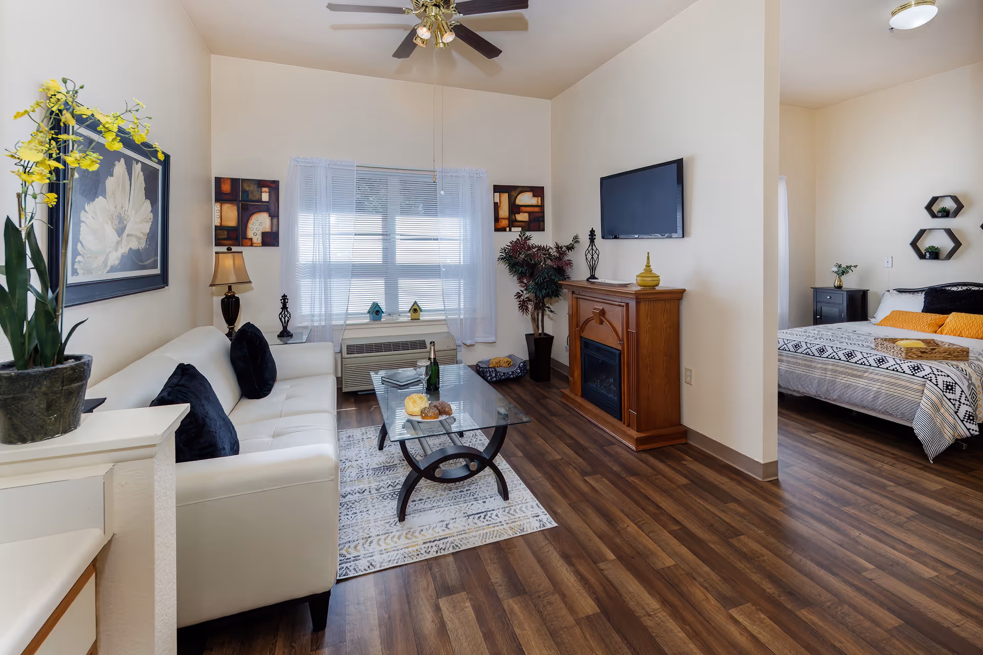 A bright and cozy living room with a white sofa adorned with dark cushions, a glass coffee table on a patterned rug, and a wooden electric fireplace with a flat-screen TV mounted above it. The room features wood flooring, a ceiling fan, and a window with sheer white curtains. To the right, a bedroom with a bed dressed in patterned bedding and yellow accent pillows is partially visible.