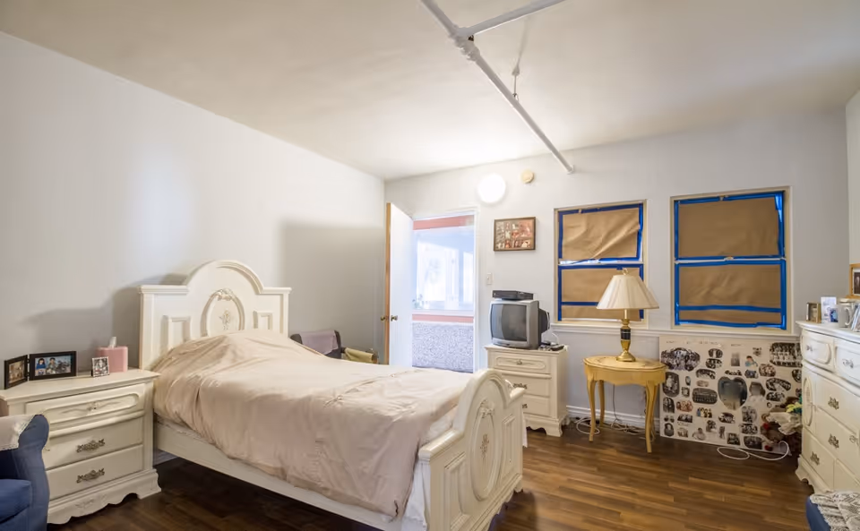 A bedroom with a white wooden bed frame and beige bedding. There are two white nightstands, one with framed photos and the other with an old-style TV and a lamp on a small round table. The windows are covered with brown paper taped with blue painter's tape. The floor is wooden, and the walls are painted white. The door is open, showing a glimpse of another room.