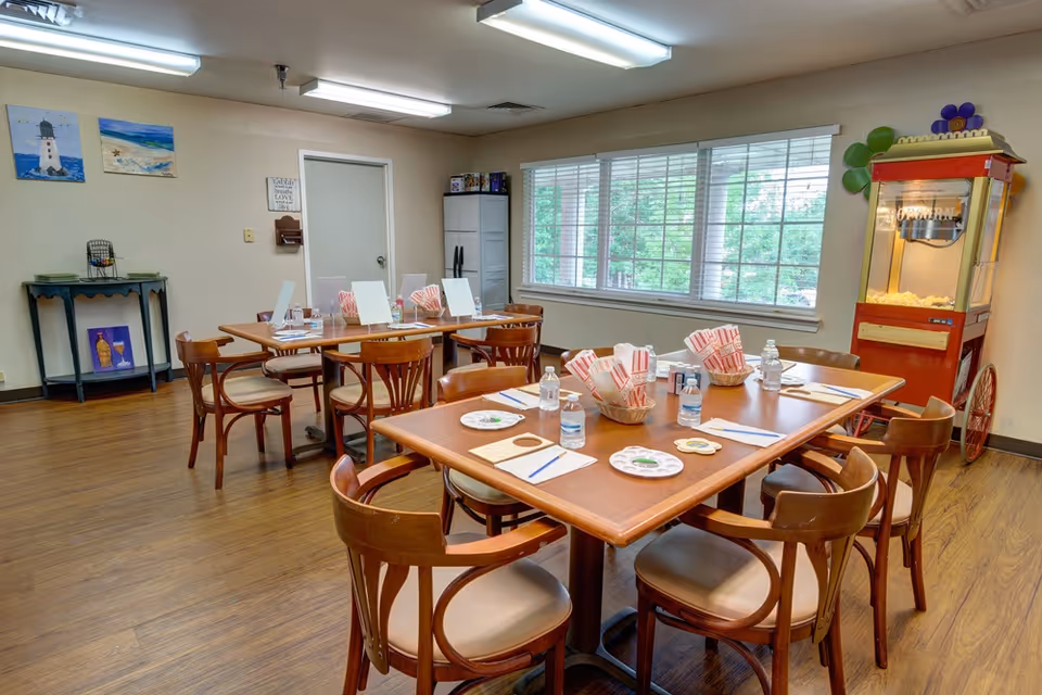 Bright communal dining/activity room with wooden tables and chairs set with water bottles, plates and popcorn baskets, large window and a popcorn machine.