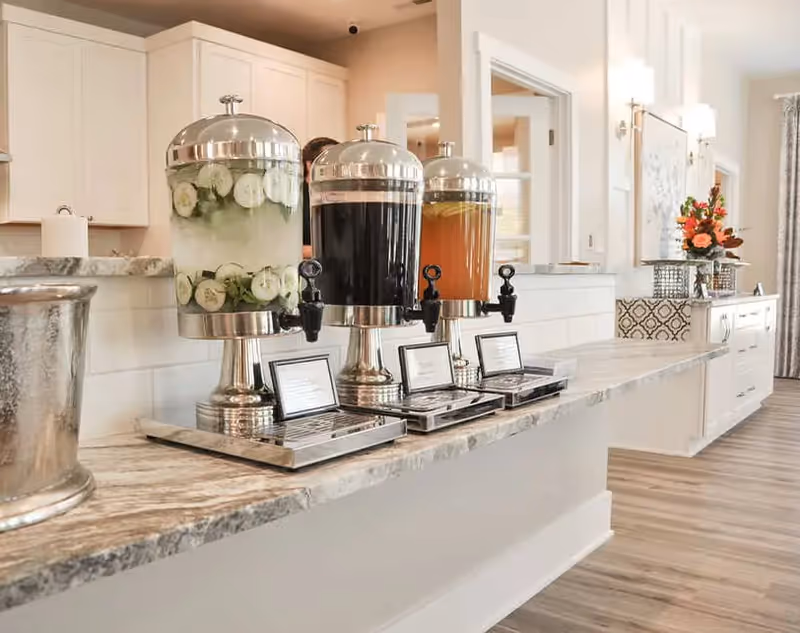 A countertop with three large beverage dispensers containing cucumber water, a dark drink, and an orange drink. The dispensers are silver and placed on a marble surface in a bright, modern interior space with white cabinets and wooden flooring. There are small framed labels in front of each dispenser and a floral arrangement in the background.