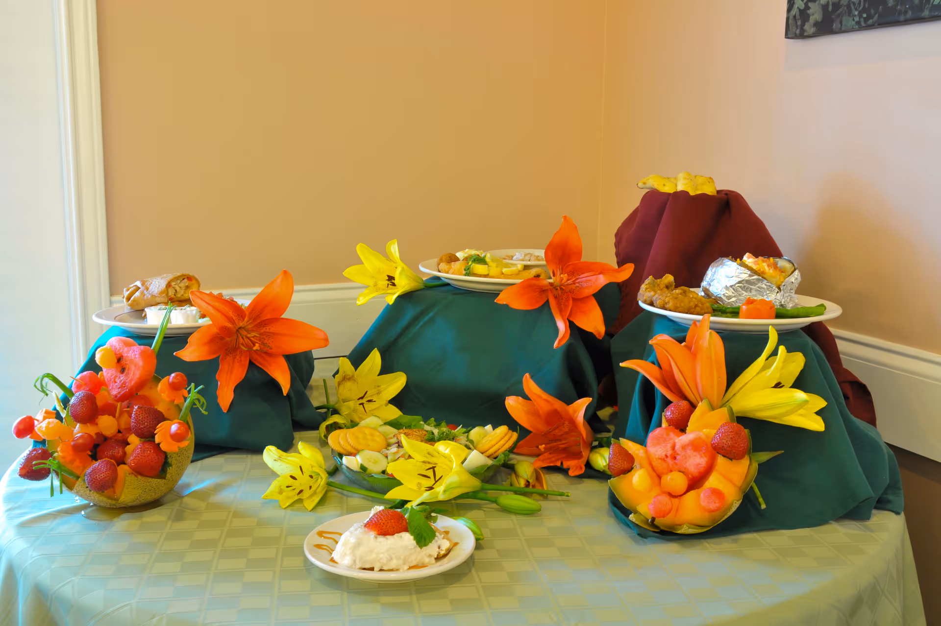 A table display featuring plates of food and decorative arrangements of colorful flowers and fruit carvings, including strawberries, melon, and other fruits, set against a beige wall.