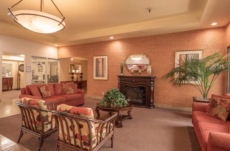 A cozy senior living facility common area with red upholstered sofas and patterned armchairs arranged around a wooden coffee table with a green plant. The room features a decorative fireplace with a mirror above it, framed artwork on the walls, and a large potted plant in the corner. Warm lighting and neutral tones create a welcoming atmosphere.