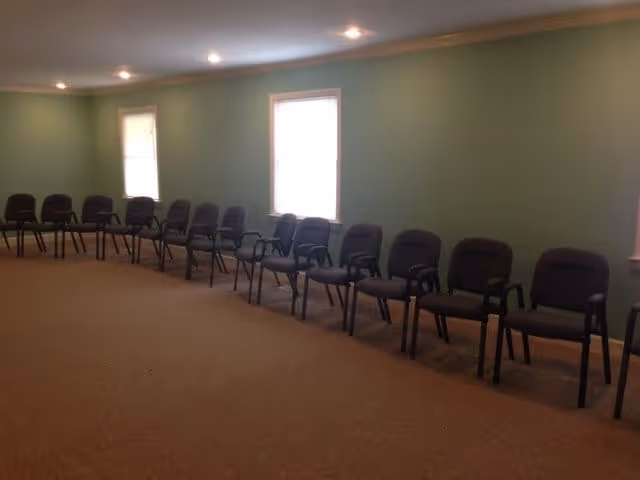 Long interior room with a row of empty cushioned chairs lined against a light green wall under recessed ceiling lights.