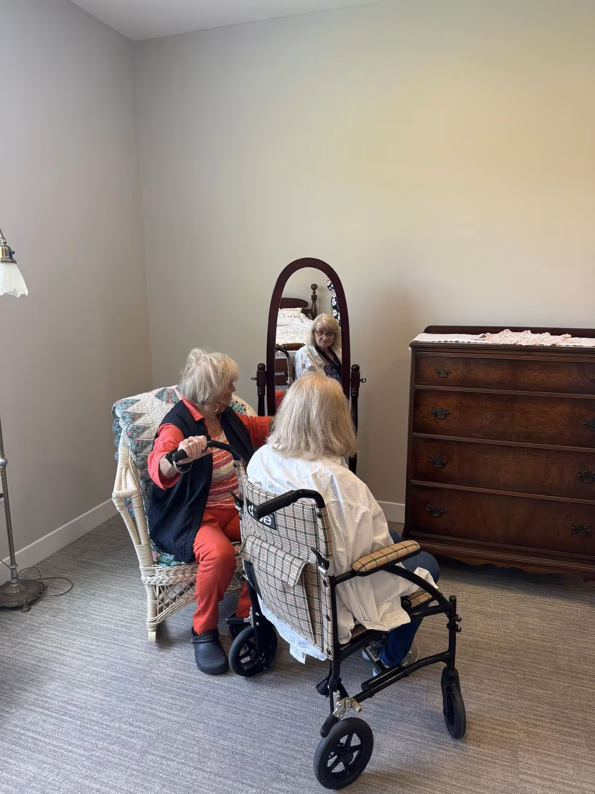 Two elderly women in a room; one is sitting in a wheelchair facing a standing mirror, and the other is seated in a wicker chair beside her. The room has a wooden dresser, a floor lamp, and neutral-colored walls and carpet.