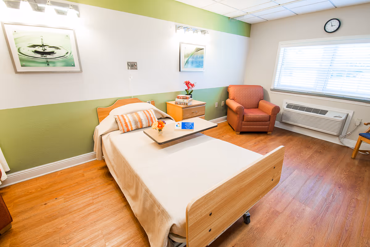 A bright and clean senior living bedroom with a single hospital-style bed, a small bedside table with flowers and books, an orange armchair, a window with blinds, and two framed pictures on the wall. The room has wooden flooring and a green and white painted wall.