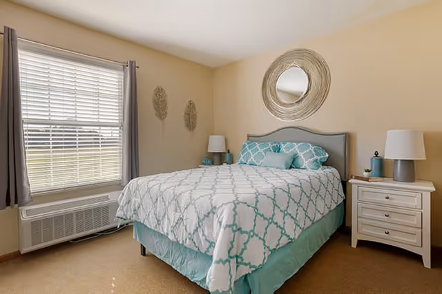 Sunlit bedroom with a double bed in teal and white patterned bedding, two nightstands with lamps, a round wall mirror, and a window with blinds.