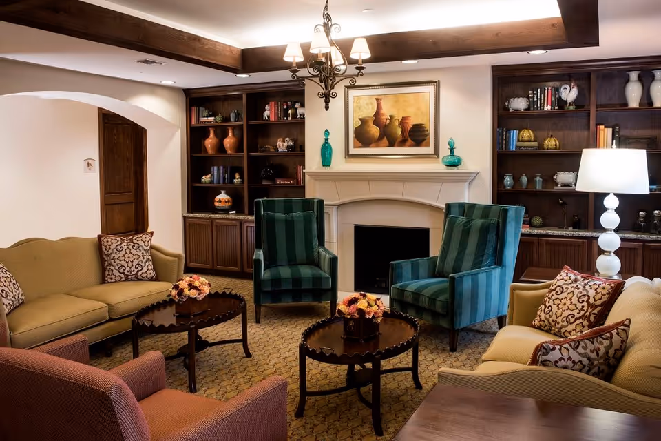 A cozy living room with two green striped armchairs, two beige sofas with patterned cushions, and a red armchair arranged around two dark wooden coffee tables with flower arrangements. The room features a fireplace with a painting of pottery above it, built-in wooden shelves with decorative items and books, a white lamp on a side table, and exposed wooden ceiling beams.