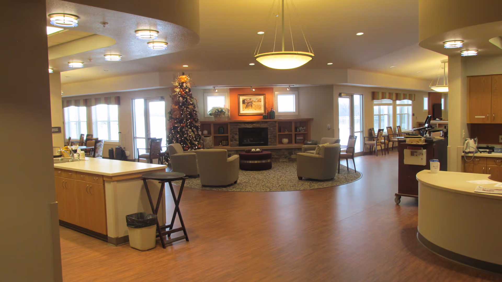 A spacious and well-lit common area in a senior living facility featuring a Christmas tree decorated with lights and ornaments near a fireplace with a framed picture above it. The room has several armchairs arranged around a circular rug, wooden flooring, and multiple tables and chairs near large windows letting in natural light. The area also includes a reception desk and cabinetry with various items on the counters.